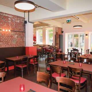 Großzügiger Restaurantbereich mit Holztischen, roten Sitzpolstern, beleuchteter Backsteinwand und großen Fenstern zur Straße