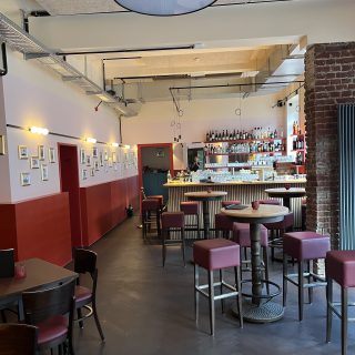 Großzügiger Barbereich mit runden Stehtischen, roten Hockern, Backsteinwand und beleuchtetem Tresen im Hintergrund