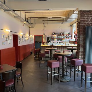 Moderner Barraum mit Stehtischen, roten Hockern, Backsteinwand und zentralem Tresen mit Spirituosenregalen