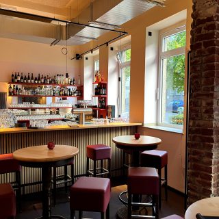 Modern gestaltete Bar mit runden Stehtischen, roten Hockern, Spirituosenregalen und großen Fenstern mit Blick ins Grüne