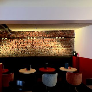 Gemütlicher Sitzbereich mit runden Tischen, roten und grauen Stühlen sowie beleuchteter Backsteinwand im Hintergrund