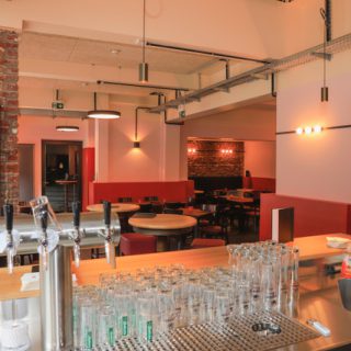 Moderner Gastraum mit runden Tischen, roter Wandgestaltung, Zapfanlage und beleuchtetem Barbereich mit Backsteinwand