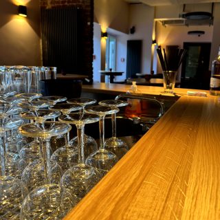 Moderne Bar mit Holztheke, gestapelten Weingläsern und warm beleuchtetem Innenraum mit Backsteinwand